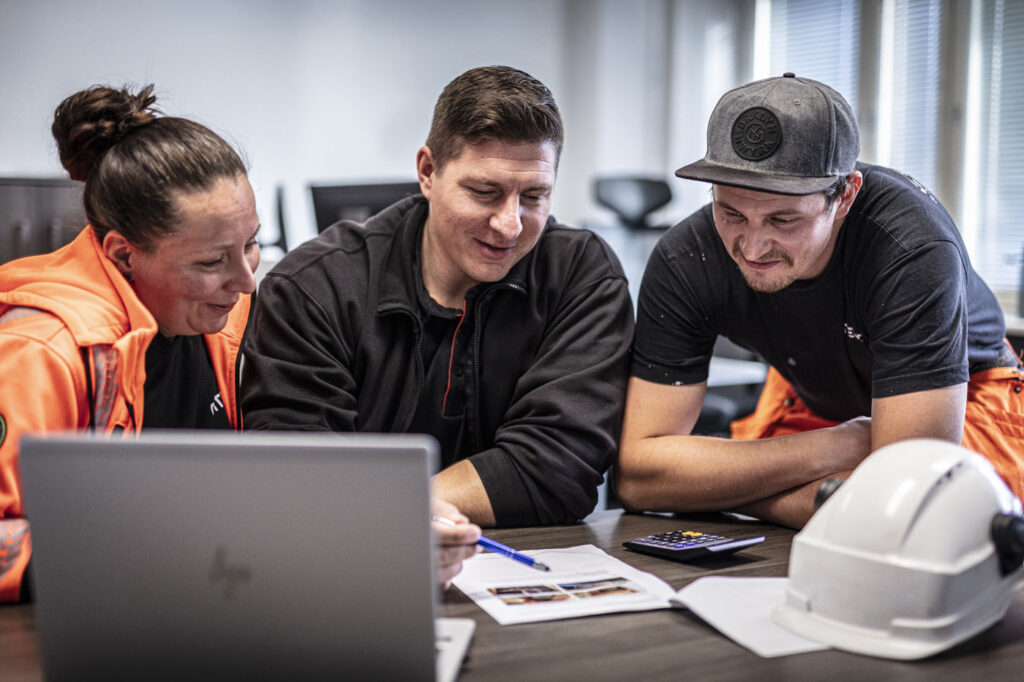 Tre personer ser på dokumenter og laptop ved bord med hjelm.