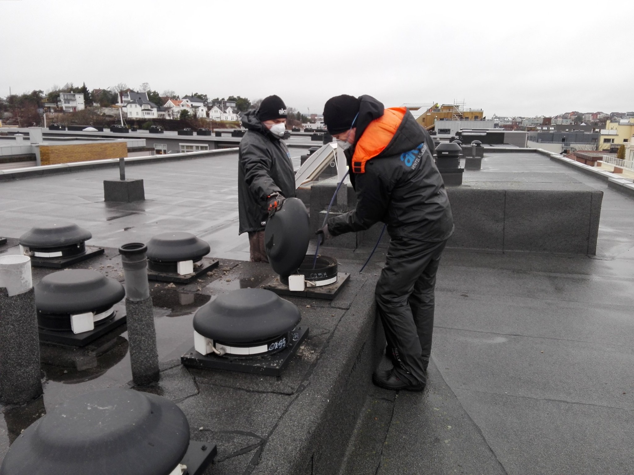 To personer arbeider med ventilasjonshetter på flatt tak i overskyet vær.